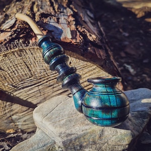 May include: A blue and green wooden smoking pipe with a curved stem. The pipe is sitting on a rock with a log in the background.