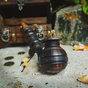 May include: A wooden smoking pipe with a dark brown finish and a round bowl. The pipe is sitting on a white sandy surface with seashells and gold coins scattered around it.