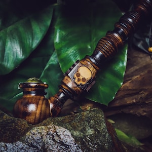 Könnte beinhalten: Eine Holzpfeife mit einem Schädelmotiv an der Seite. Die Pfeife liegt auf einem Stein mit grünen Blättern im Hintergrund.