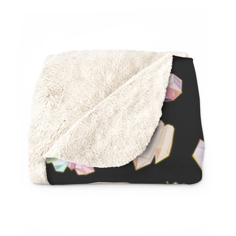 Może przedstawiać: Miękki, pluszowy koc z czarnym tłem i powtarzającym się wzorem kolorowych, geometrycznych kryształ&oacute;w. Koc jest złożony na p&oacute;ł, pokazując białą, puszystą stronę spodnią.