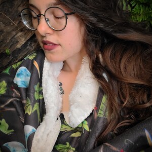 May include: A woman wearing round glasses and a black and green patterned fleece robe with a white sherpa collar. The robe has a floral design with a moon and stars pattern.