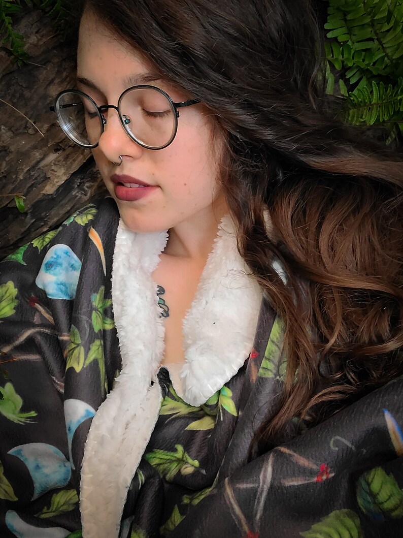 May include: A woman wearing round glasses and a black and green patterned fleece robe with a white sherpa collar. The robe has a floral design with a moon and stars pattern.
