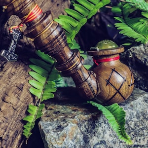 May include: A handcrafted wooden pipe with a decorative bowl and stem. The pipe features a silver pendant of a hammer and is accented with red and yellow details. The pipe is surrounded by green ferns and rests on a stone surface.