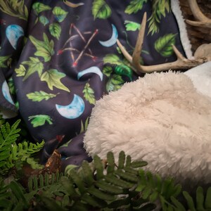 May include: A soft, white faux fur throw blanket with a dark blue and green patterned throw blanket underneath. The patterned blanket features crescent moons, leaves, and sticks.
