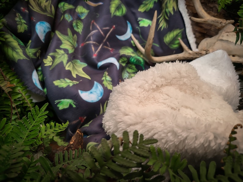 May include: A soft, white faux fur throw blanket with a dark blue and green patterned throw blanket underneath. The patterned blanket features crescent moons, leaves, and sticks.