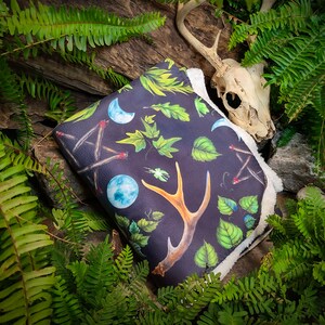 May include: A black fleece blanket with a white sherpa lining features a repeating pattern of antlers, leaves, moons, and matches. The blanket is lying on a bed of green ferns and a deer skull.