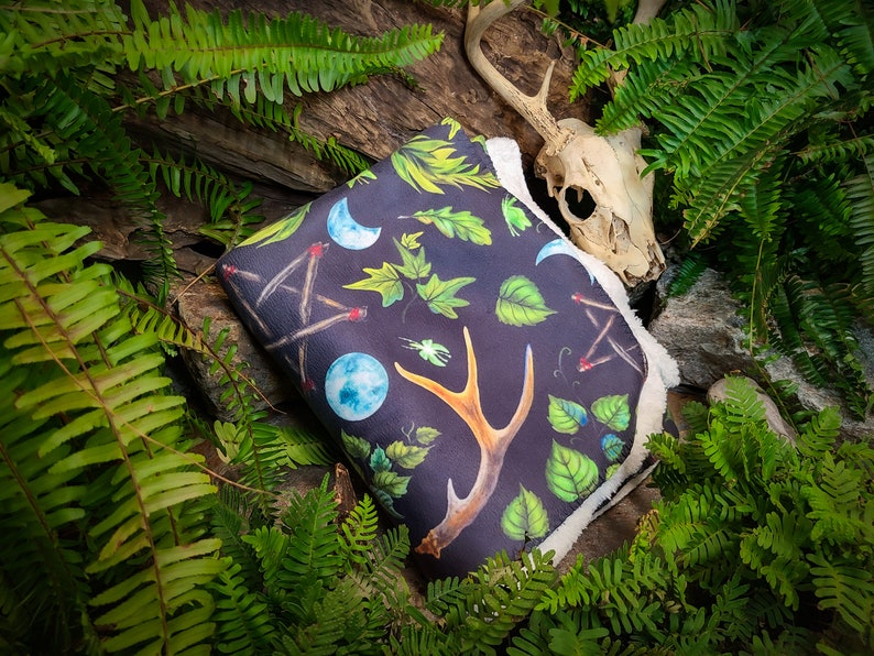 May include: A black fleece blanket with a white sherpa lining features a repeating pattern of antlers, leaves, moons, and matches. The blanket is lying on a bed of green ferns and a deer skull.