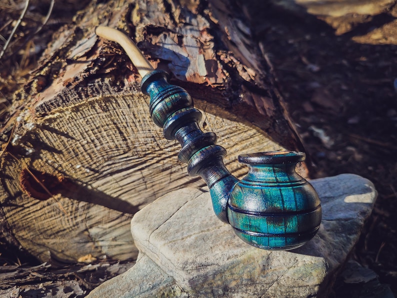 May include: A blue and green wooden smoking pipe with a curved stem. The pipe is sitting on a rock with a log in the background.
