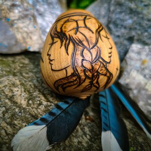 Op de afbeelding: Een houten, eivormig decoratief item met geëtste profielen van twee vrouwen. Het lichtbruine hout is versierd met donkerbruine geëtste details. Blauwe en witte veren zijn aan de onderkant bevestigd, waardoor een uniek, artistiek stuk ontstaat.