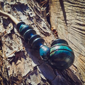 May include: A wooden pipe with a blue and green iridescent finish. The pipe is made of multiple sections and has a bowl at the end. The pipe is resting on a tree trunk.