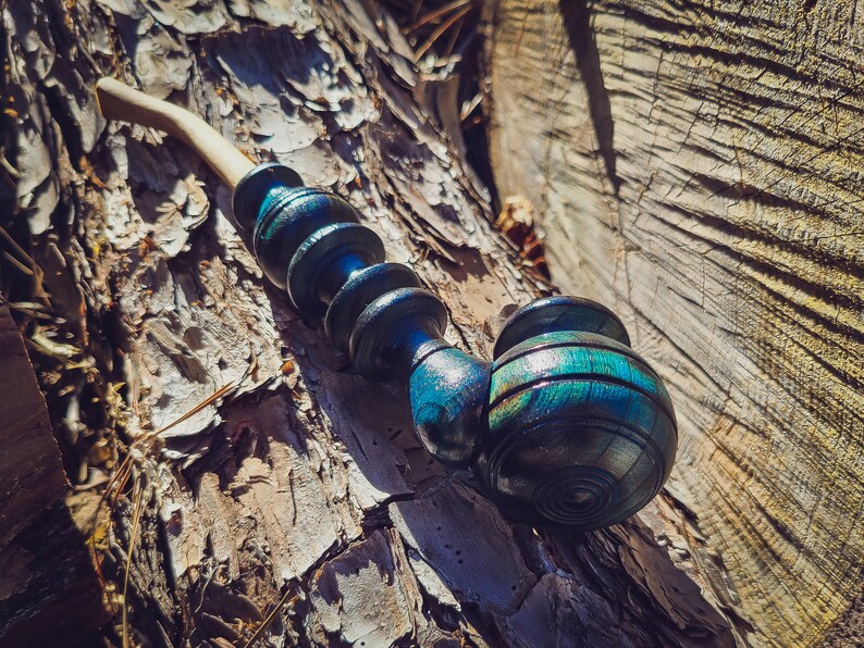 May include: A wooden pipe with a blue and green iridescent finish. The pipe is made of multiple sections and has a bowl at the end. The pipe is resting on a tree trunk.