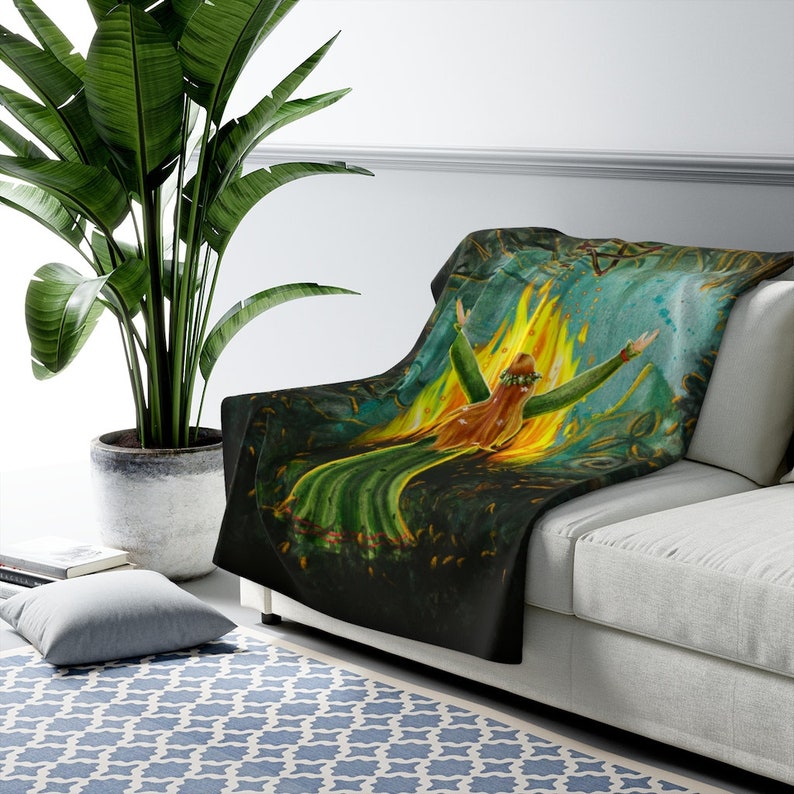 May include: A black throw blanket with a colorful illustration of a woman standing in a fire. The woman is wearing a green dress and a crown of leaves. The background is a dark forest with a blue sky.