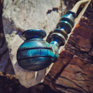 May include: A blue and green wooden pipe with a round bowl. The pipe is resting on a piece of tree bark.