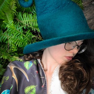 May include: A woman wearing a teal green felt witch hat with a curled brim and a black and green patterned shawl. She is wearing glasses and has brown hair.