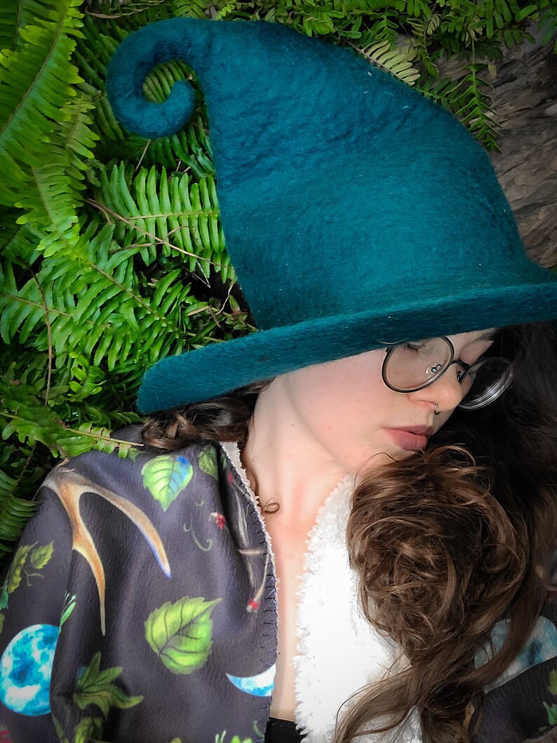 May include: A woman wearing a teal green felt witch hat with a curled brim and a black and green patterned shawl. She is wearing glasses and has brown hair.