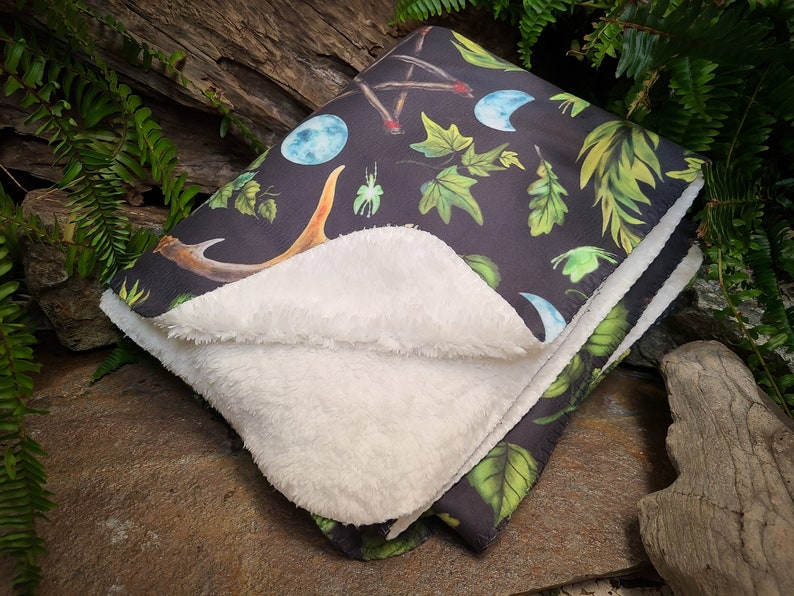 May include: A soft, black and white fleece blanket with a dark green, black, and white print of moons, antlers, leaves, and a pentagram. The blanket is folded in half and lying on a rock.