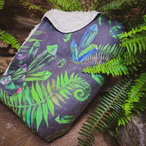 May include: A soft, plush throw blanket with a dark background featuring a repeating pattern of green ferns and colorful crystals. The blanket has a white sherpa lining.