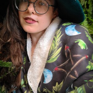 May include: A woman wearing a green hat and round glasses is wrapped in a black and green blanket with a white fleece lining. The blanket has a moon and leaf pattern.