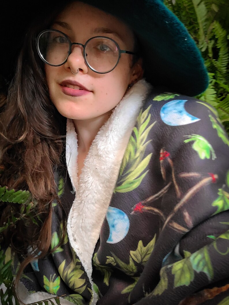 May include: A woman wearing a green hat and round glasses is wrapped in a black and green blanket with a white fleece lining. The blanket has a moon and leaf pattern.
