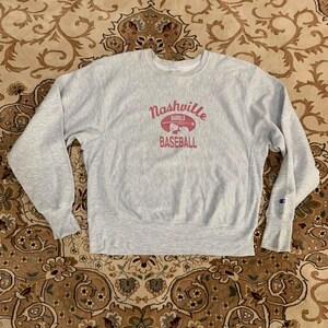 May include: A light grey Champion sweatshirt featuring the text "Nashville Barrels Baseball" in pink. The sweatshirt has long sleeves and a crew neck. A small Champion logo is visible on the left sleeve.