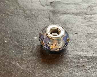 Op de afbeelding: Een bolvormige bedel van helder glas met ingesloten kleine blauwe bloemetjes en dunne donkere vezels. De bedel heeft een centrale kern van sterling zilver met het keurmerk 925 gegraveerd op het gepolijste metalen oppervlak.