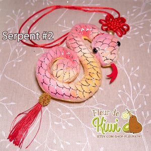 Op de afbeelding: Een roze en gele pluche slang met een rode kwast en een rode knoop. De slang is opgerold en heeft een glinsterende afwerking. De tekst "Serpent #2" is zichtbaar in de afbeelding.