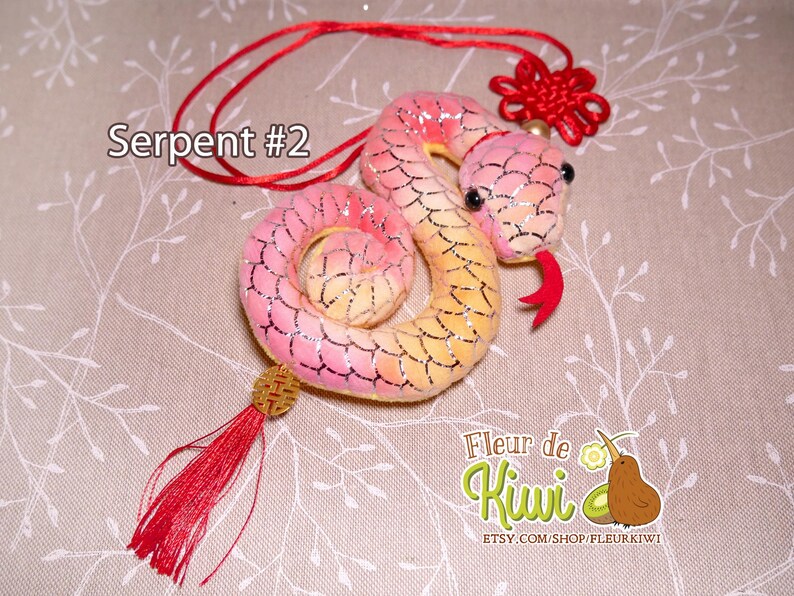 Op de afbeelding: Een roze en gele pluche slang met een rode kwast en een rode knoop. De slang is opgerold en heeft een glinsterende afwerking. De tekst "Serpent #2" is zichtbaar in de afbeelding.