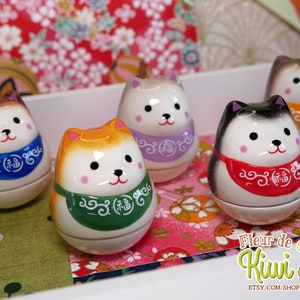 May include: Five ceramic dog-shaped figurines with colourful scarves and painted details. The figurines are traditional Japanese "daruma" dolls, which are lucky charms. The dolls are arranged on a table with a floral patterned tablecloth.