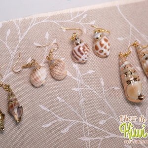 May include: A collection of seashell earrings with gold-colored hooks. The earrings feature various shell shapes and sizes, with natural colors and gold accents. The earrings are displayed on a beige fabric with a floral pattern.