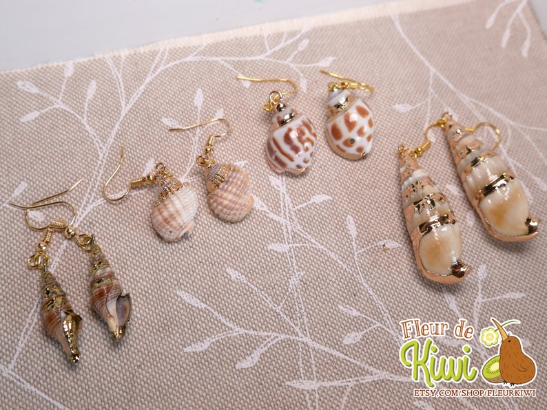 May include: A collection of seashell earrings with gold-colored hooks. The earrings feature various shell shapes and sizes, with natural colors and gold accents. The earrings are displayed on a beige fabric with a floral pattern.