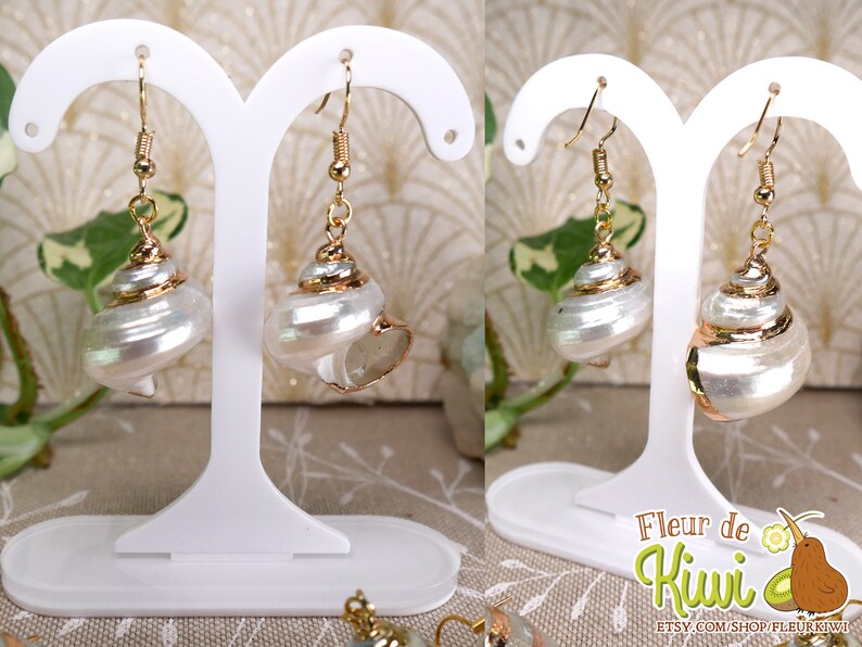 May include: A pair of seashell earrings with gold-colored accents. The earrings feature a pearlescent white shell design with a gold-colored rim and hook. The earrings are displayed on a white stand.