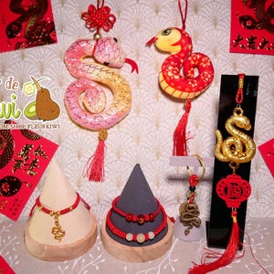 Op de afbeelding: Een set Chinese nieuwjaarsdecoraties met een rode en gouden slang, een rode en gouden draak en een gouden slangenhanger. De decoraties zijn op een witte achtergrond met rode en gouden enveloppen en een bord met de tekst "Fleur de Kiwi".