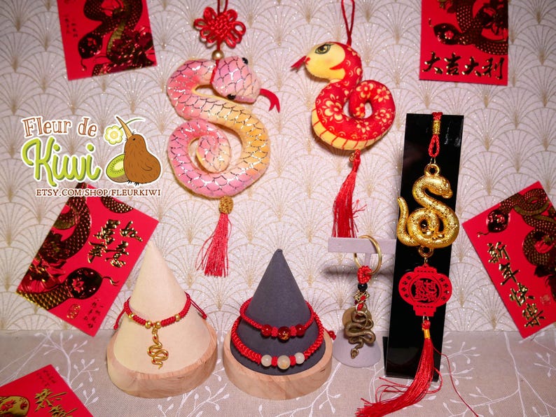 Op de afbeelding: Een set Chinese nieuwjaarsdecoraties met een rode en gouden slang, een rode en gouden draak en een gouden slangenhanger. De decoraties zijn op een witte achtergrond met rode en gouden enveloppen en een bord met de tekst "Fleur de Kiwi".