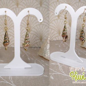 May include: Gold-toned seashell earrings with spiral details hang from gold hooks. The earrings are displayed on a white earring stand. The seashells have a mix of brown, tan, and gold tones. The earrings are a beach-themed accessory.