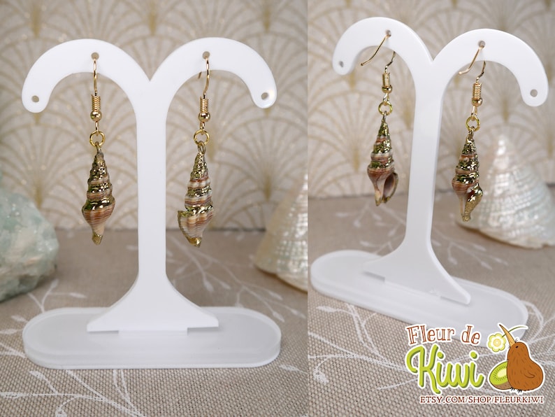 May include: Gold-toned seashell earrings with spiral details hang from gold hooks. The earrings are displayed on a white earring stand. The seashells have a mix of brown, tan, and gold tones. The earrings are a beach-themed accessory.