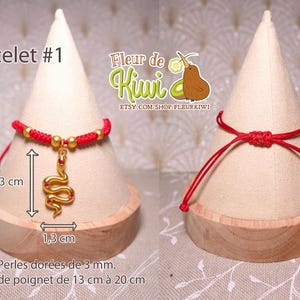 Op de afbeelding: Rood koord armband met een gouden slang bedel. Het armband is 3 cm lang en de bedel is 1,3 cm lang. Het armband is gemaakt met 3 mm gouden kralen. Het armband wordt getoond op een houten display stand.