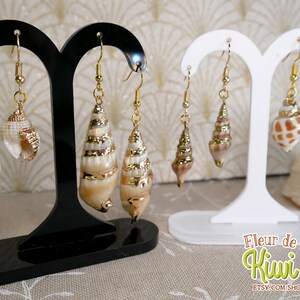 May include: Earrings featuring natural seashells with gold-colored accents. The earrings are displayed on black and white stands. The seashells have a variety of shapes and sizes, with gold-colored metal caps and hooks. The earrings are a beach-themed accessory.