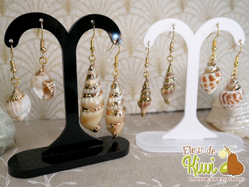 May include: Earrings featuring natural seashells with gold-colored accents. The earrings are displayed on black and white stands. The seashells have a variety of shapes and sizes, with gold-colored metal caps and hooks. The earrings are a beach-themed accessory.