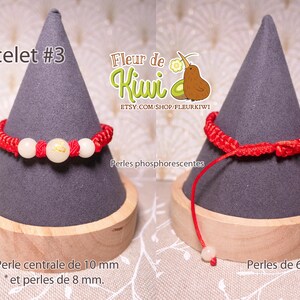 Op de afbeelding: Rood koord armband met witte kralen. De centrale kraal is 10 mm en de andere kralen zijn 8 mm. Het armband wordt getoond op een grijze standaard met de tekst "Bracelet #3" en "Fleur de Kiwi" op de standaard.