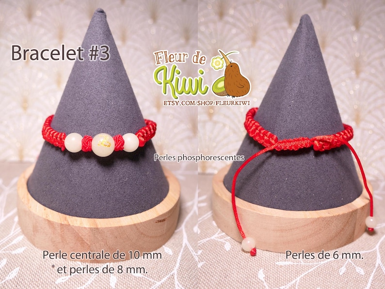 Op de afbeelding: Rood koord armband met witte kralen. De centrale kraal is 10 mm en de andere kralen zijn 8 mm. Het armband wordt getoond op een grijze standaard met de tekst "Bracelet #3" en "Fleur de Kiwi" op de standaard.