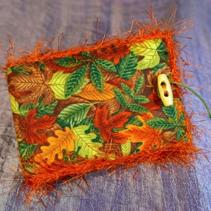 May include: A rectangular fabric item with a fall leaves pattern in shades of brown, orange, and green. It has an orange fringe border and a wooden button closure with a green string.