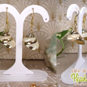 May include: A pair of seashell earrings with gold accents. The earrings are displayed on a white stand. The shells are cream-colored with gold-colored edges and details. The earrings have gold-colored hooks.