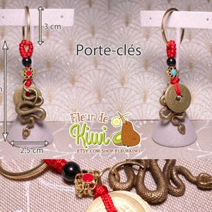 Op de afbeelding: Een gouden slangenhanger met een munt en rode kralen hangt aan een gouden sleutelhanger. De hanger is bevestigd aan een grijze basis en is 7 cm lang. De hanger is 2,5 cm breed. De hanger is 3 cm hoog. De hanger is een slang met een munt op zijn kop. De munt heeft een Chinees karakter erop. De hanger is een sleutelhanger.