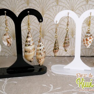 May include: Earrings featuring natural seashells with gold-colored accents. The earrings are displayed on black and white earring stands. The seashells vary in shape and size, with some having a spiral design. The earrings are suspended from gold-colored hooks.