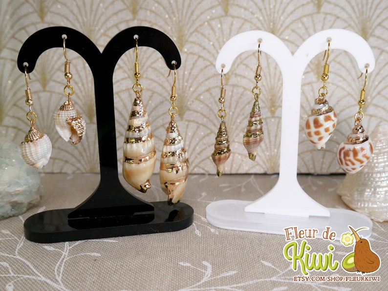 May include: Earrings featuring natural seashells with gold-colored accents. The earrings are displayed on black and white earring stands. The seashells vary in shape and size, with some having a spiral design. The earrings are suspended from gold-colored hooks.