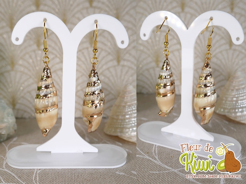 May include: A pair of seashell earrings with gold-colored accents. The earrings are displayed on a white stand. The seashells have a spiral design and are a light beige color.