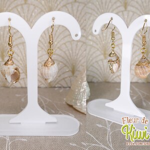 May include: Two seashell earrings with gold-colored accents are displayed on a white earring stand. The earrings feature natural-colored shells with gold-colored caps and hooks. The earrings are showcased against a soft background.