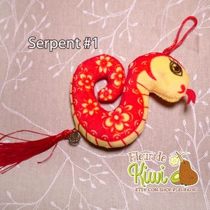 Op de afbeelding: Rood en geel pluche slangen speelgoed met een bloemenpatroon. De slang is opgerold en heeft een klein rood kwastje eraan bevestigd. Het speelgoed is gelabeld met "Serpent #1" en "Fleur de Kiwi".