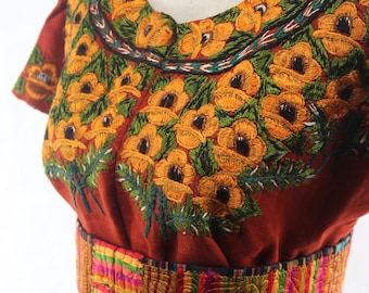 Może przedstawiać: Bordowana bluzka w kolorze rdzawo-brązowym z kwiatowym wzorem w odcieniach żółtego, zielonego i czarnego. Bluzka ma szeroki, kolorowy, tkany pasek.