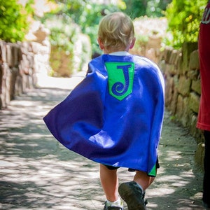 May include: A young child wearing a blue cape with a green and blue patch that has the letter "J" on it. The child is walking on a paved path.